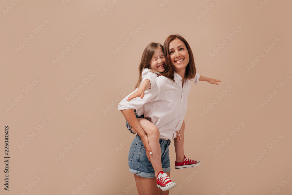 Inspired young woman without make-up spending time with daughter, carrying her piggyback over isolated background.