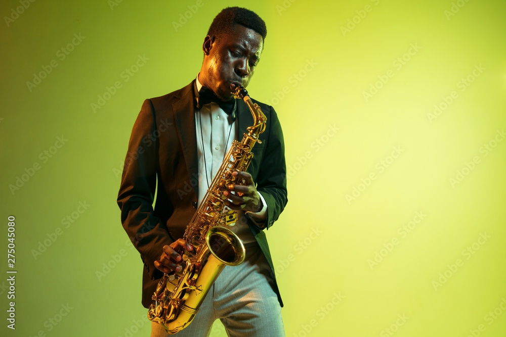 Naklejka premium Young african-american jazz musician playing the saxophone on gradient yellow-green studio background. Concept of music, hobby, festival. Joyful attractive guy improvising. Colorful portrait of artist