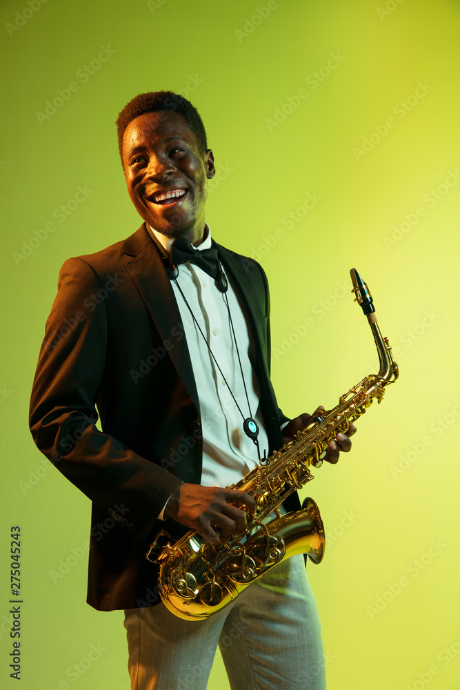 Fototapeta premium Young african-american jazz musician playing the saxophone on gradient yellow-green studio background. Concept of music, hobby, festival. Joyful attractive guy improvising. Colorful portrait of artist
