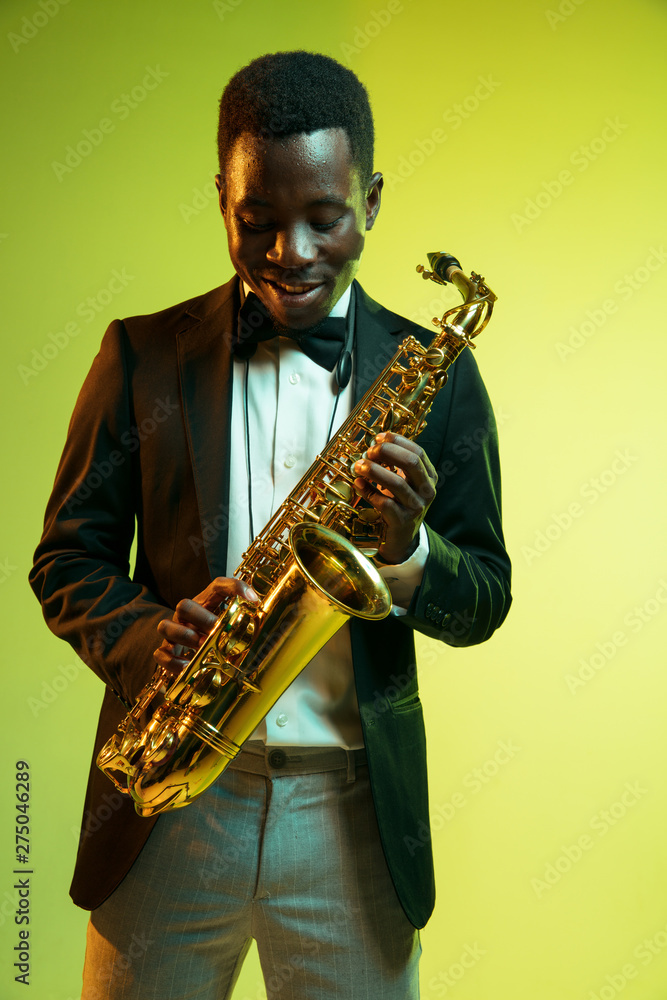 Naklejka premium Young african-american jazz musician playing the saxophone on gradient yellow-green studio background. Concept of music, hobby, festival. Joyful attractive guy improvising. Colorful portrait of artist