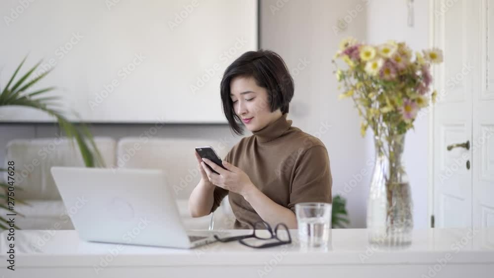 Pan shot of smiling asian business woman with short haircut scrolling the phone sitting to table with laptop. Glass of water on table and a vase with flowers in white home office Pan shot of smiling asian business woman with short haircut scrolling the phone sitting to table with laptop. Glass of water on table and a vase with flowers in white home office