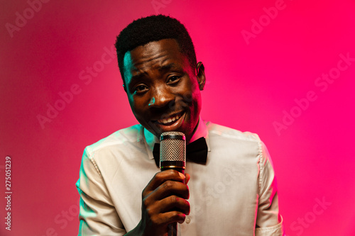 Young african-american jazz musician singing a song on gradient pink-red background in neon light. Concept of music, hobby, festival, open-air. Joyful guy improvising. Colorful retro portrait of