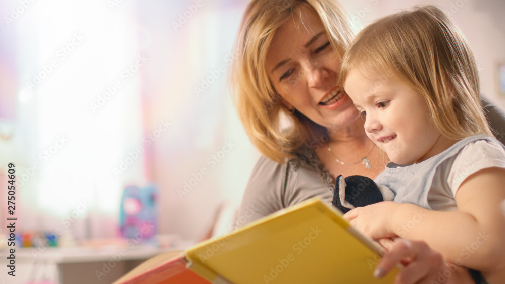 Close-up Shot of a Beautiful Young Mother and Her Cute Little Daughter Read Children's Book Together. Children's Room is Pink and Full of Toys. Close-up Shot of a Beautiful Young Mother and Her Cute Little Daughter Read Children's Book Together. Children's Room is Pink and Full of Toys.