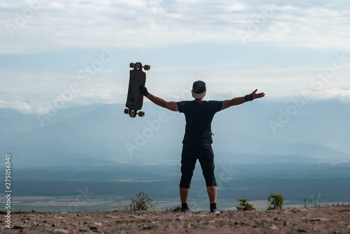 A young man in a cap with a Board in his hands stands on a cliff high in the mountains against the sky, a journey on a skateboard,