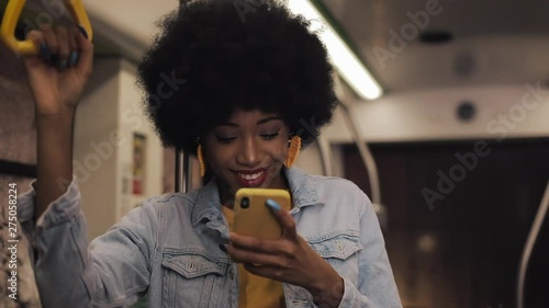 Attractive young african american woman holds the handrail and using smartphone at public transport. Night time.