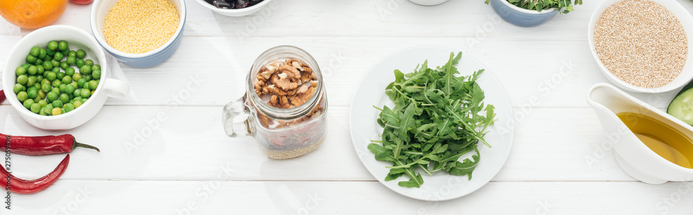 top view of glass jar with nuts near plate with green arugula on wooden white table, panoramic shot top view of glass jar with nuts near plate with green arugula on wooden white table, panoramic shot