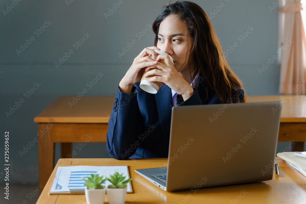 Female office worker wears dark blue business suit works with laptop while drinking coffee in smiling face in the office. Adult business woman working and drinking coffee in relaxing manner. Female office worker wears dark blue business suit works with laptop while drinking coffee in smiling face in the office. Adult business woman working and drinking coffee in relaxing manner.
