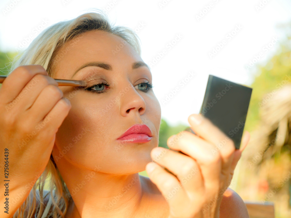 young beautiful and attractive blond woman with blue eyes retouching makeup with brush applying eyeshadow holding small mirror in female beauty and fashion young beautiful and attractive blond woman with blue eyes retouching makeup with brush applying eyeshadow holding small mirror in female beauty and fashion