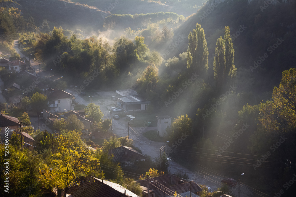 Aerial view of beautiful foggy village between mountains in Lovech, Bulgaria. Misty sunrise view of city district surrounded by rock mountain. Sun beams between trees