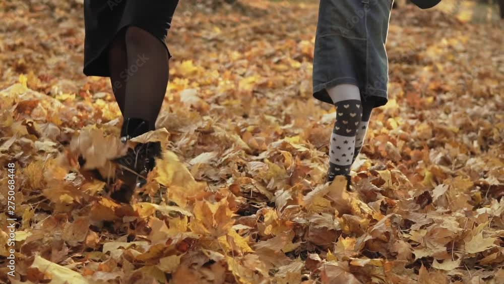 Daughter with her mother kicking autumn leaves by boots in park. Slow motion. Low angle. Close up view of legs Daughter with her mother kicking autumn leaves by boots in park. Slow motion. Low angle. Close up view of legs