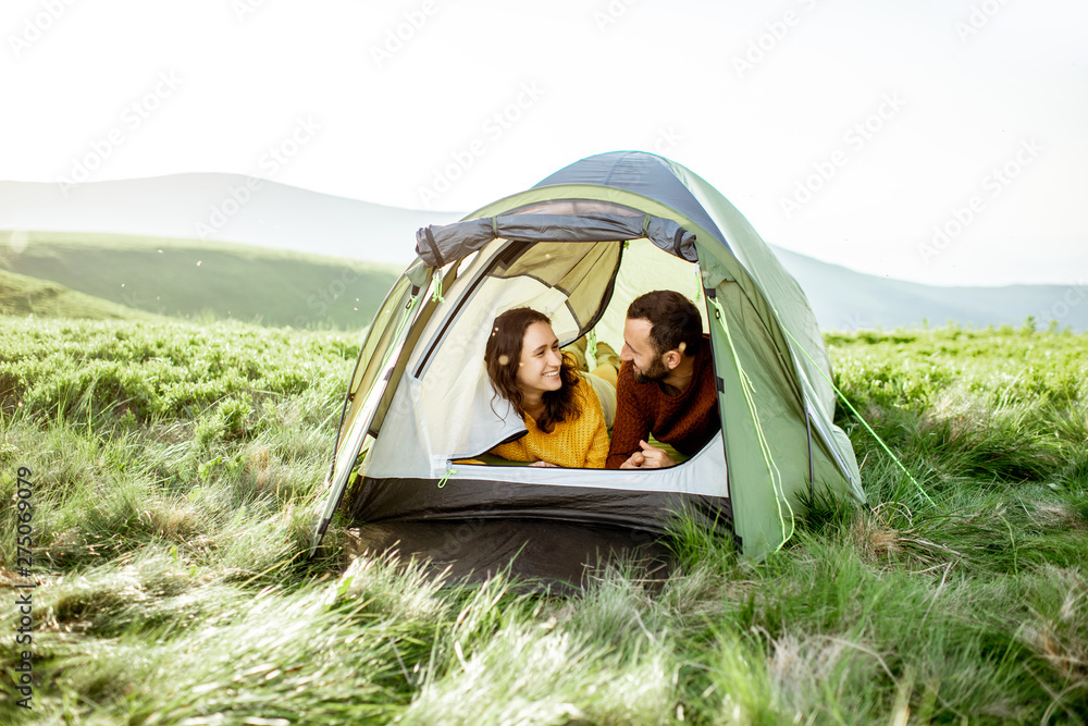 Young lovely couple in bright sweaters enjoying nature, lying in the tent while traveling high in the mountains