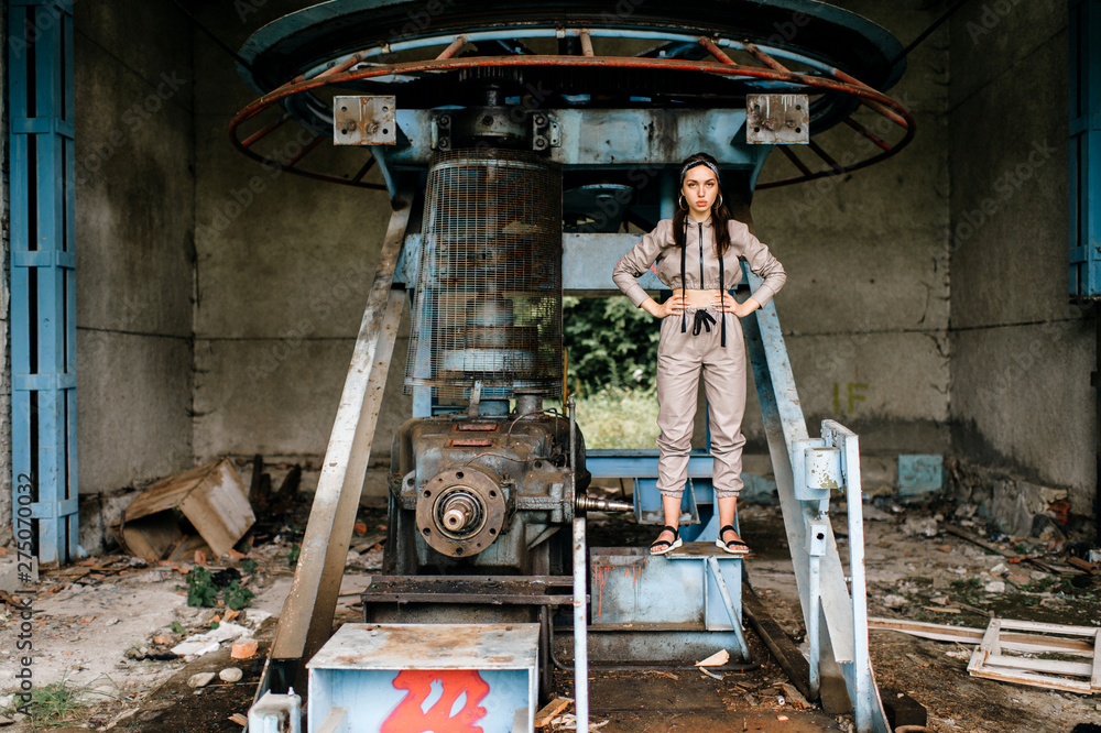 Beautiful young stylish girl posing at abandoned factory outdoor with an old broken engine.