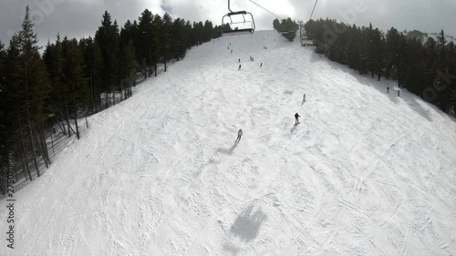 Skiers and snowboarders going downhill on a mountain slope at a ski resort with snowy forest, ski lift and mountains in the background under a clear sky, POV slow motion