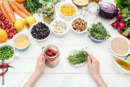cropped view of woman holding glass jar with salad and plate with herbs on wooden white table