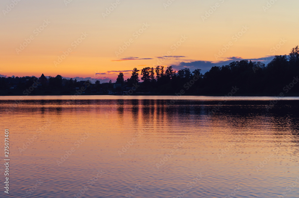 Golden and purple sky reflected in the water of a lake. Black silhouettes of trees at dusk. Enjoy peaceful time and atmosphere of tranquility in the evening twilight.