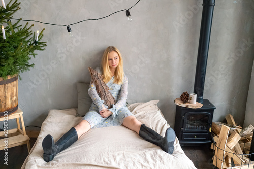 Schilderij op canvas Stylish girl in rubber boots siiting on bed and posing with decorative log in her hands in rural room with christmas tree