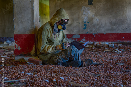 Man in respirator gas mask, steampunk glasses, hood and camo pants sits on the floor inside of abandoned building and reading book. Cyberpunk postapocalypse fantasy horror scene, or air pollution © evgenzz