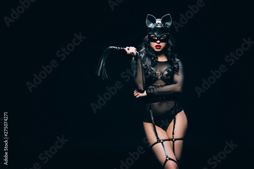 sexy young brunette woman in bdsm costume and mask with leather flogging whip isolated on black