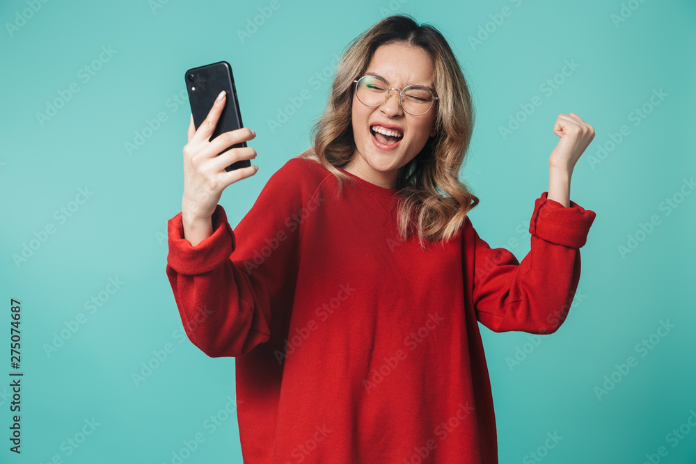 Emotional young woman posing isolated over blue wall background using mobile phone make winner gesture. Emotional young woman posing isolated over blue wall background using mobile phone make winner gesture.