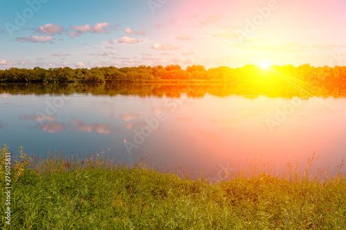 Pink orange summer evening sky. Sun rays and clouds reflecting in water. Meadow and forest on banks. © geniousha
