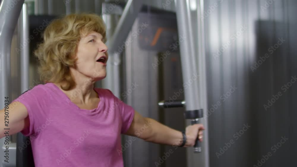 Old woman sitting on exercise machine and pumping arms muscles while senior man standing nearby showing her video lesson on tablet Old woman sitting on exercise machine and pumping arms muscles while senior man standing nearby showing her video lesson on tablet