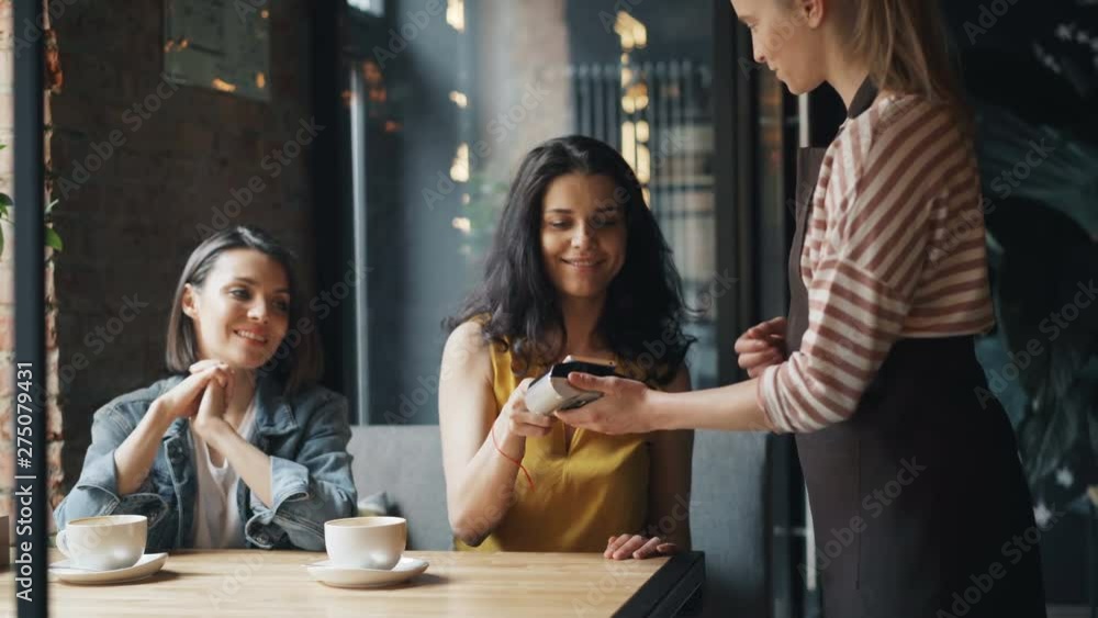 Young woman paying for lunch in cafe using smartphone talking to waitress in apron smiling enjoying modern technology. Customers and business concept. Young woman paying for lunch in cafe using smartphone talking to waitress in apron smiling enjoying modern technology. Customers and business concept.