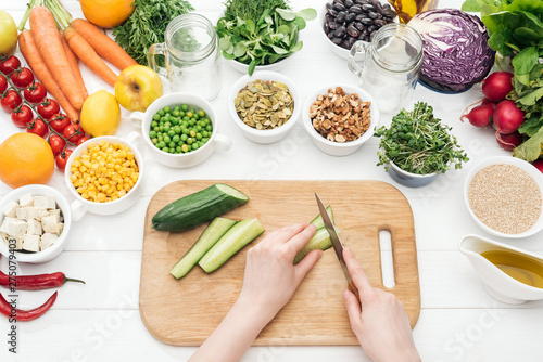 cropped view of woman cutting cucumber on wooden white table