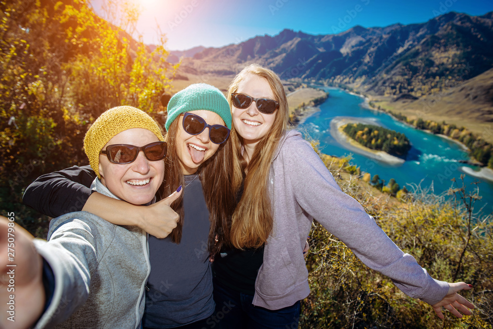 European women in sunglasses laugh doing selfie against mountain background. Young female hikers pose against amazing small island on the mountain river. Outdoor recreation concept.