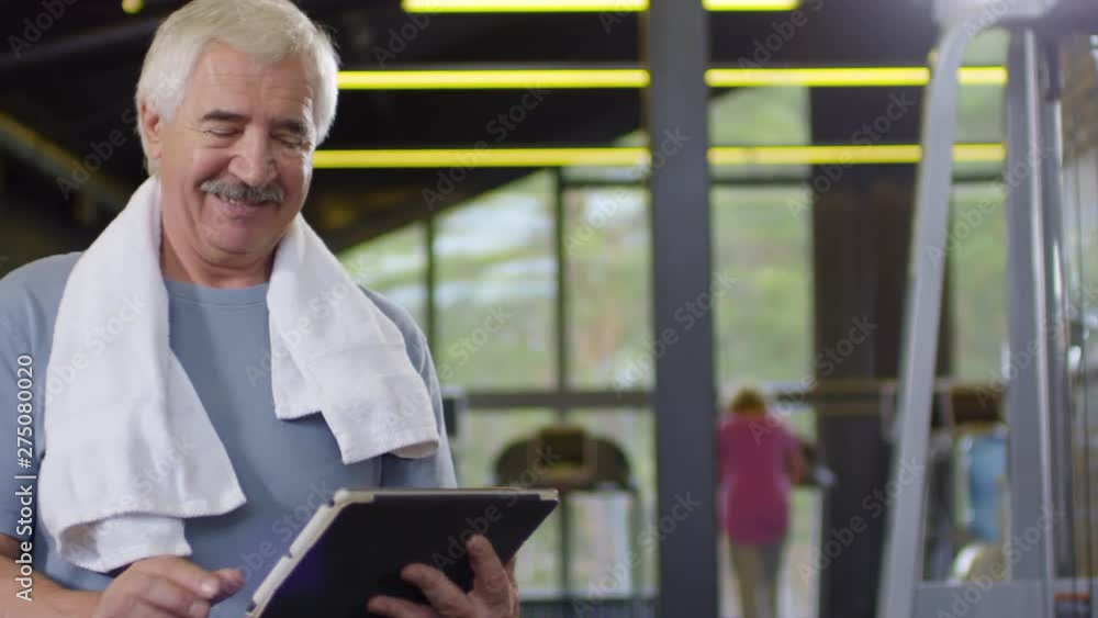 Tracking left of senior Caucasian man standing in gym after training with tablet in his hands and swiping Tracking left of senior Caucasian man standing in gym after training with tablet in his hands and swiping
