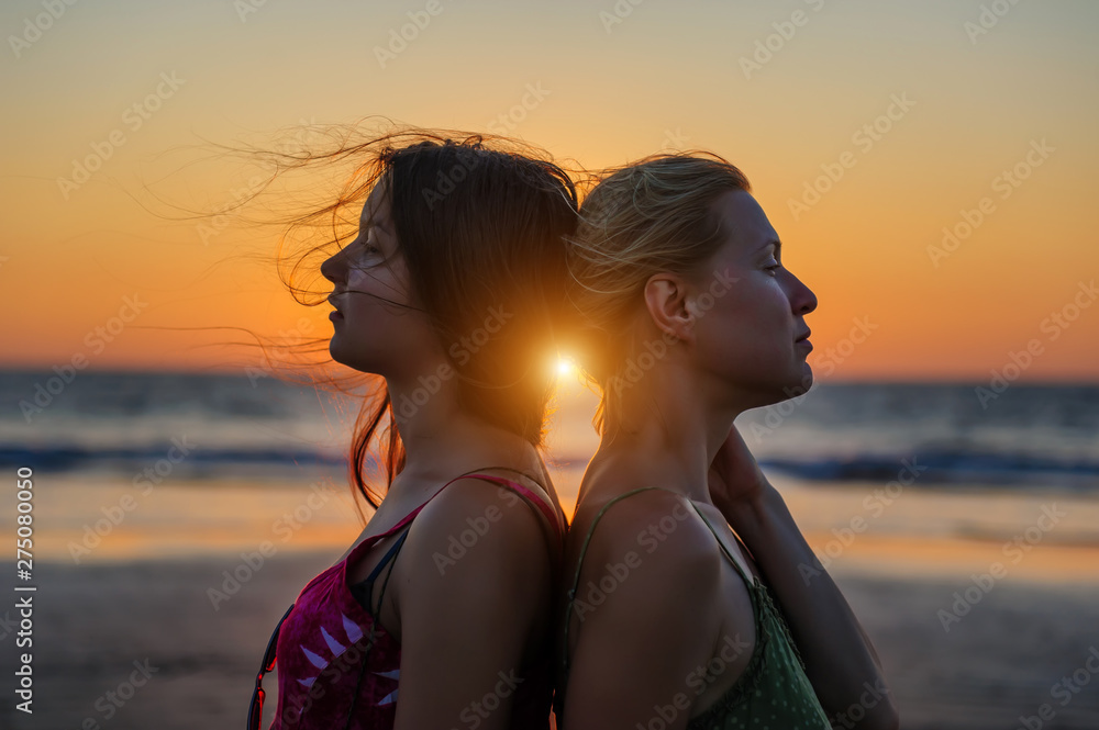 Blonde and brunette girlfriends are clinging back to each other against sunset over the sea. Happy lesbian european couple is resting on tropical beach. LGBT concept.