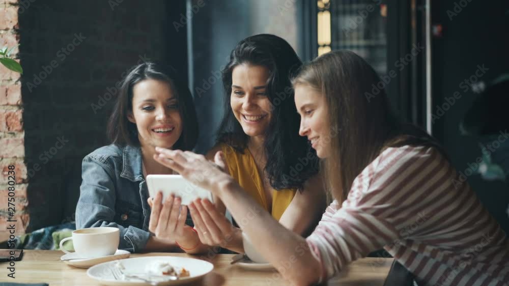 Joyful girls happy friends are looking at smartphone screen swiping smiling having fun in cafe enjoying media content. Modern technology and friendship concept. Joyful girls happy friends are looking at smartphone screen swiping smiling having fun in cafe enjoying media content. Modern technology and friendship concept.