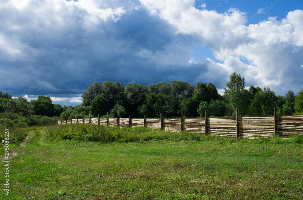 Wooden fence set in a forest area next to a rural road in the background blue sky with clouds