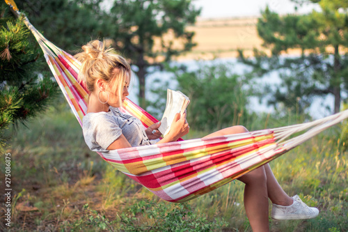 young attractive caucasian woman reading book in hammock in the forest, lake on the background