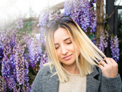 Blonde girl with round glasses on the background of wisteria. Spring portrait of pretty woman.