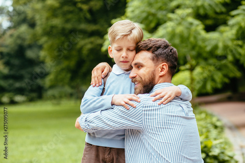 Relations father's tenderness concept. Beard handsome dad hugging his son outdoors in green park on background. Man saying goodbye to his little child before back to school.