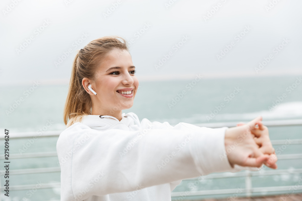 Image of young fitness woman smiling and stretching her arms while doing workout near seaside