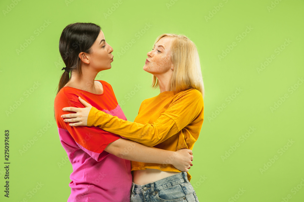 Portrait of pretty charming girls in casual outfits isolated on green studio background. Two female models as a girlfriends or lesbians. Concept of LGBT, equality, human emotions, love, relation. Portrait of pretty charming girls in casual outfits isolated on green studio background. Two female models as a girlfriends or lesbians. Concept of LGBT, equality, human emotions, love, relation.