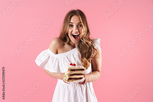 Image of attractive surprised woman wearing white dress smiling at camera and holding piece of cake with candle
