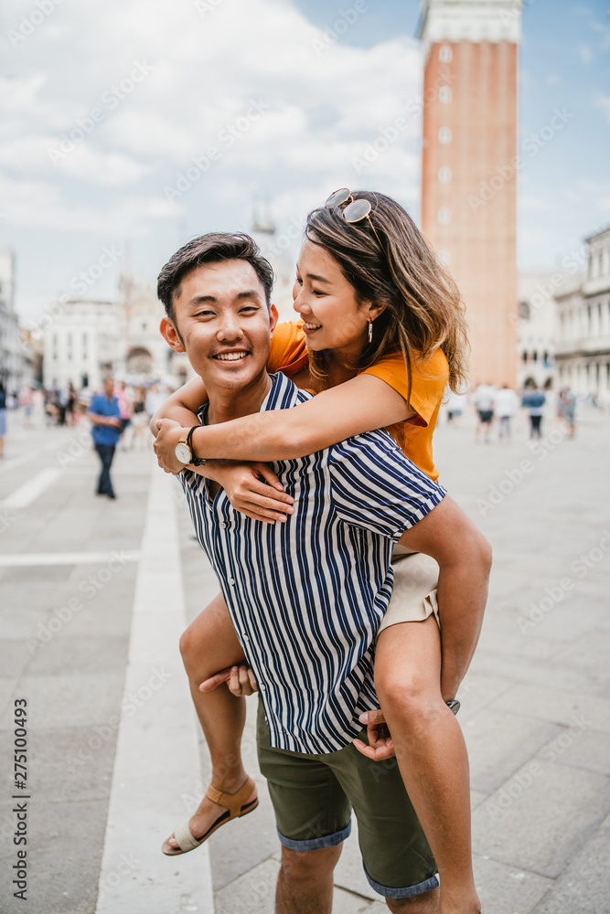 Couple of asian lovers in Piazza San Marco in Venice. The woman is on the man's shoulders as she laughs with the city's cathedral in the background