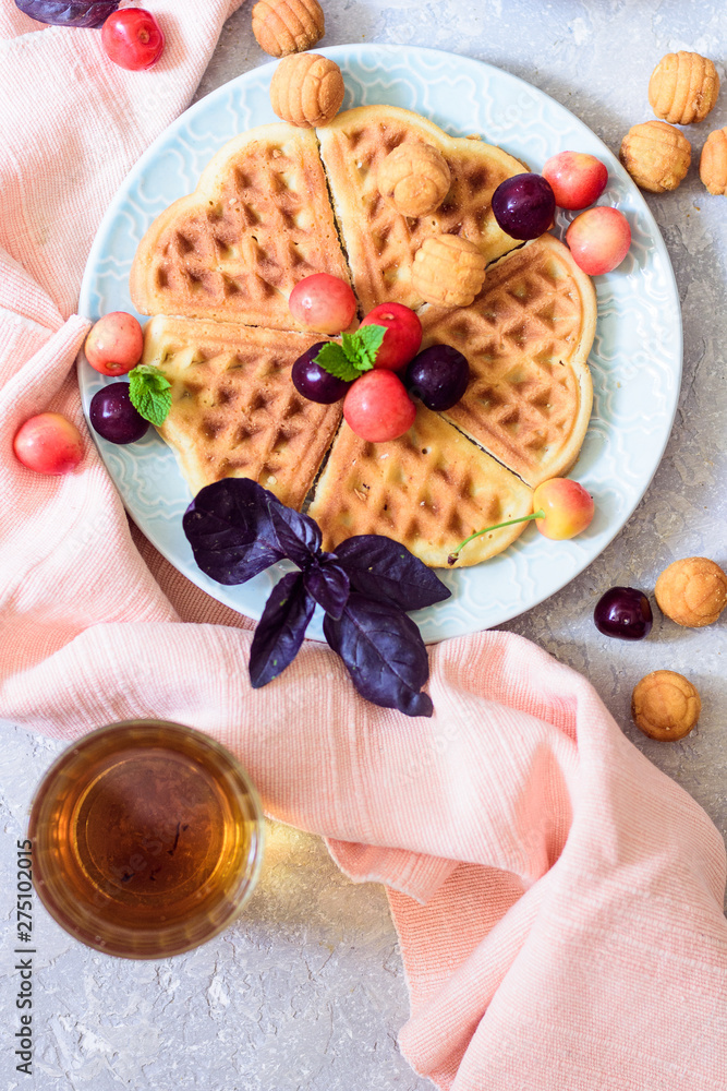 Belgian waffles on a plate with cherry and tea top view on a gray background with a pink napkin