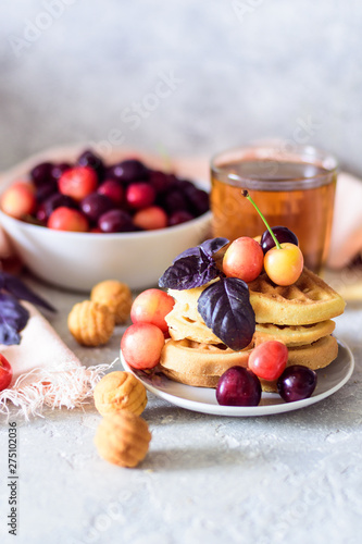 Belgian waffles on a plate with cherries and tea on a gray concrete background with a pink napkin