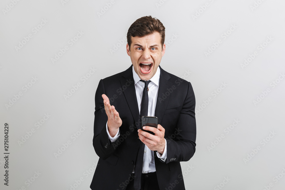 Image of angry yong businessman in formal suit typing on cellphone and screaming at camera Image of angry yong businessman in formal suit typing on cellphone and screaming at camera