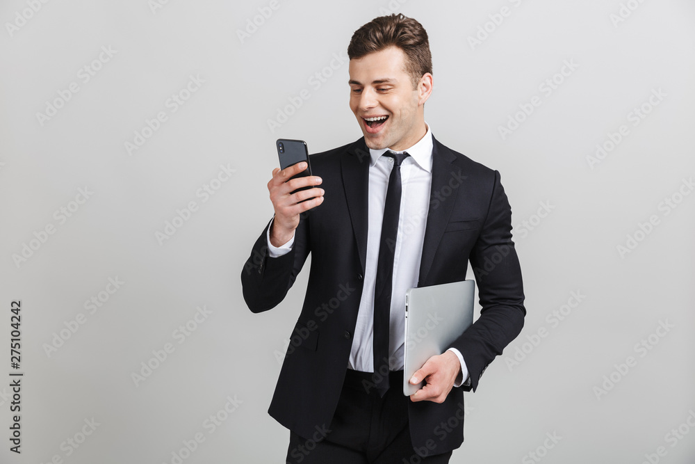 Image of confident happy businessman in formal suit typing on cellphone and holding laptop Image of confident happy businessman in formal suit typing on cellphone and holding laptop