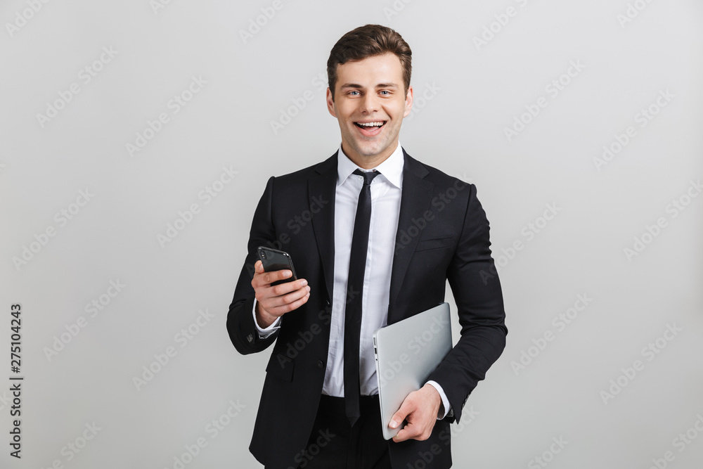 Image of young happy businessman in formal suit smiling while holding laptop and cellphone isolated Image of young happy businessman in formal suit smiling while holding laptop and cellphone isolated