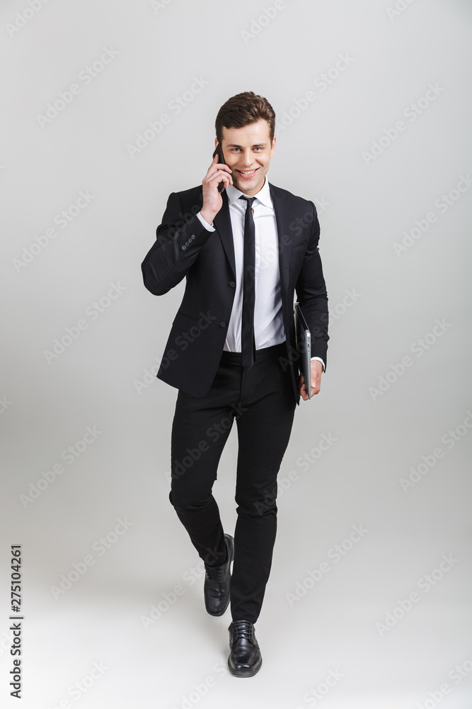 Full length image of young positive businessman in formal suit talking on cellphone and holding laptop while walking Full length image of young positive businessman in formal suit talking on cellphone and holding laptop while walking