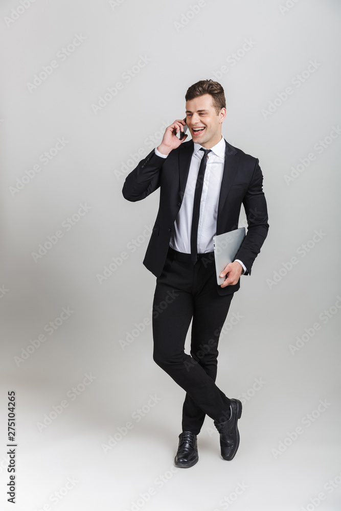 Full length image of handsome positive businessman in formal suit talking on cellphone and holding laptop Full length image of handsome positive businessman in formal suit talking on cellphone and holding laptop