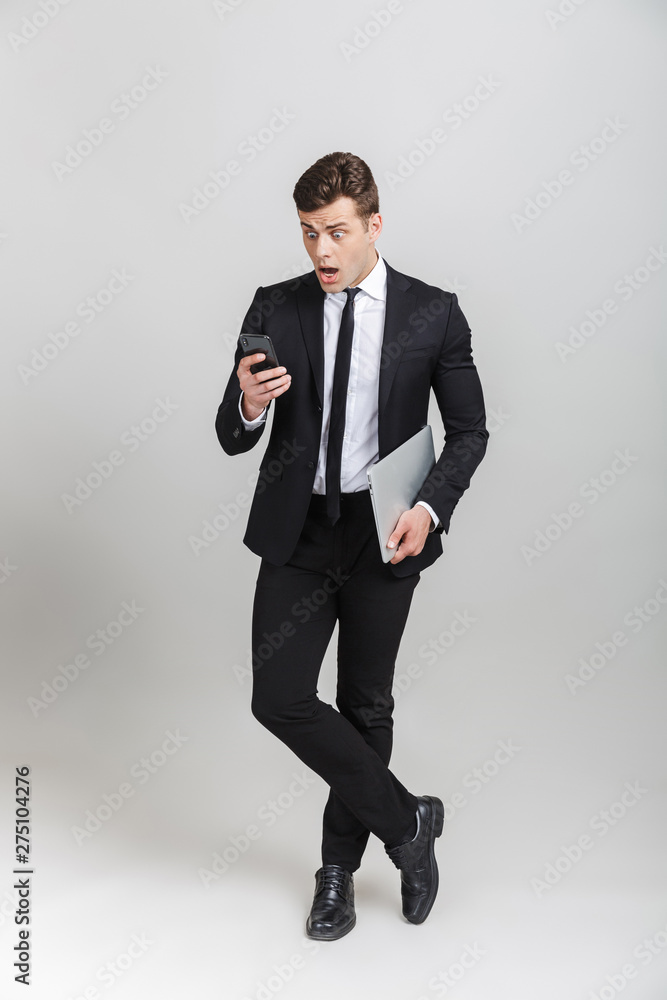 Full length image of tense businessman in formal suit looking on cellphone and holding laptop Full length image of tense businessman in formal suit looking on cellphone and holding laptop