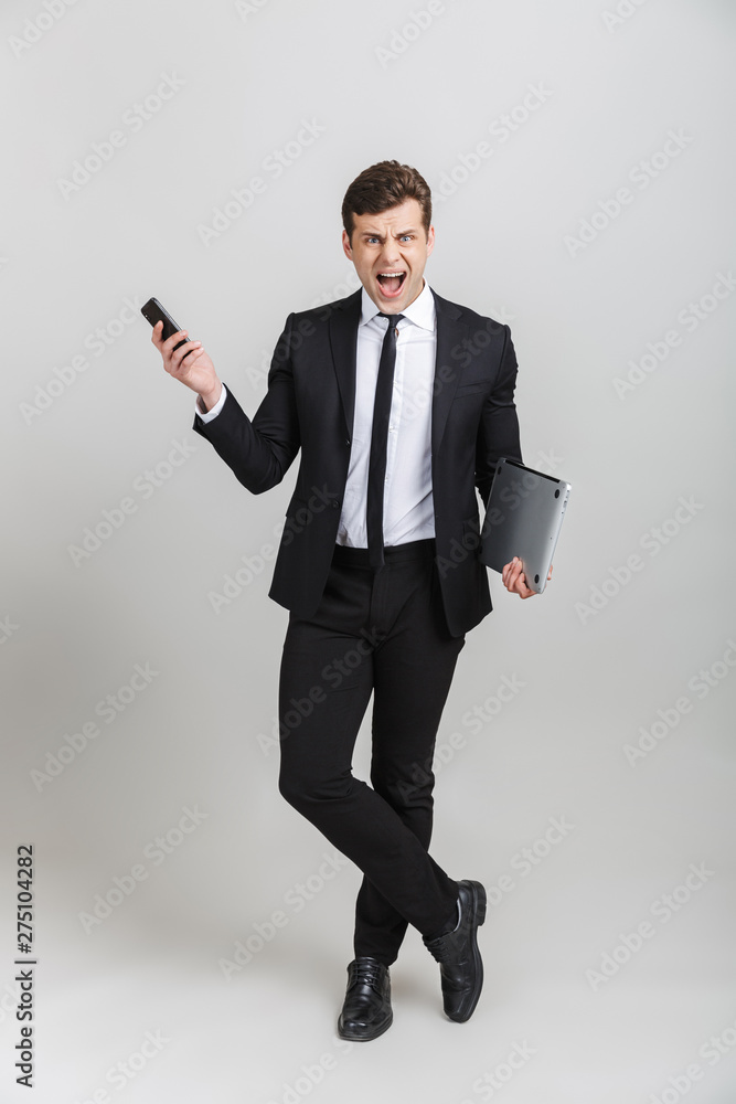 Full length image of furious businessman in formal suit screaming while holding cellphone and laptop Full length image of furious businessman in formal suit screaming while holding cellphone and laptop