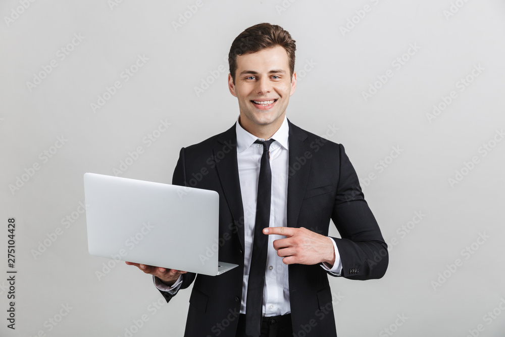 Image of attractive brunette businessman in formal suit smiling at camera while pointing finger at laptop Image of attractive brunette businessman in formal suit smiling at camera while pointing finger at laptop