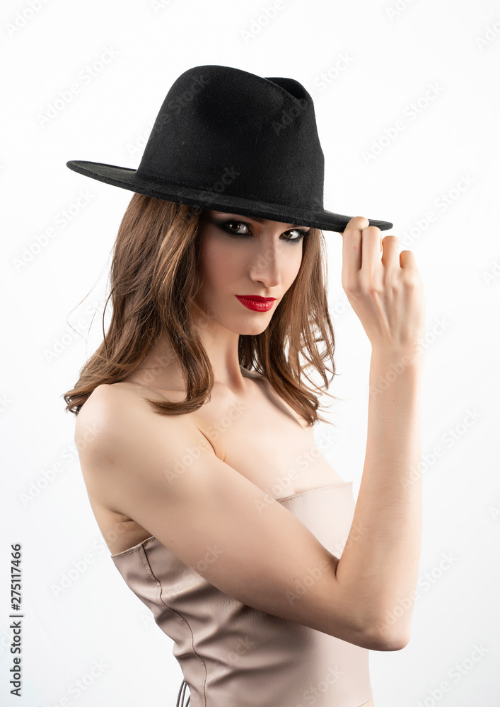 Beautiful girl model with red lips make-up and naked shoulders smiles and touches the black hat worn on her head. Isolated on white Beautiful girl model with red lips make-up and naked shoulders smiles and touches the black hat worn on her head. Isolated on white